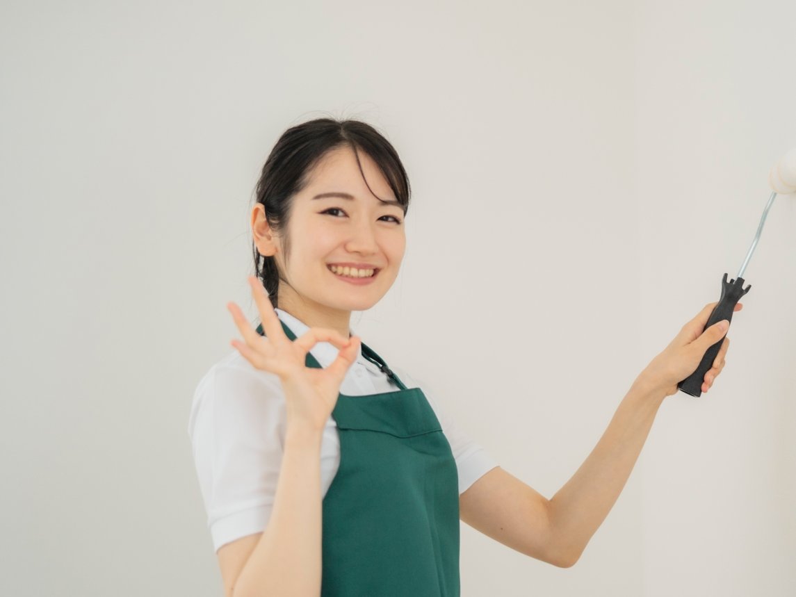 白い壁をローラーで塗装しながら、OKサインをするエプロン姿の女性